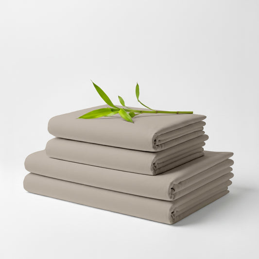 Stack of neatly folded taupe bamboo bed sheets with a green leafy twig on top against a white background.